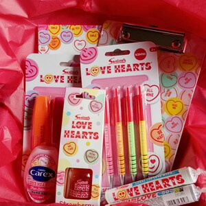 May include: A selection of Valentine's Day gifts, including Love Hearts sweets, pens, hand sanitiser, and a clipboard. The items are presented in a red gift box with red tissue paper. The packaging features heart-shaped designs.