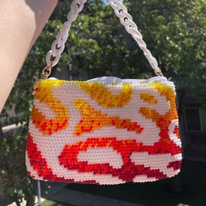 May include: A small white beaded bag with a white chain strap. The bag has a red, orange, and yellow abstract pattern.