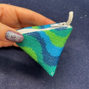 May include: A small, triangular zippered pouch made of green, blue, and white beads. The pouch is closed and held in a hand.