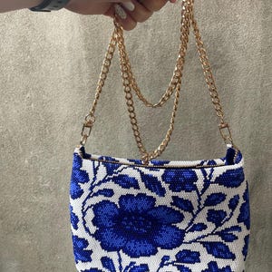 May include: A small, rectangular handbag with a blue and white floral pattern. The bag has a gold chain strap and a zipper closure. The floral design features large blue flowers and leaves against a white background. The bag is held up by a hand.
