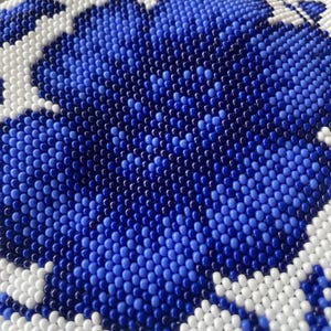 May include: Close-up of a beaded surface with a floral design. The pattern features a large blue flower with white and blue accents. The beads are small and create a textured, detailed appearance.