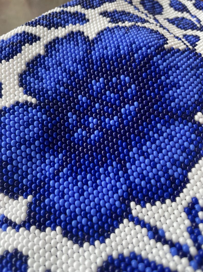 May include: Close-up of a beaded surface with a floral design. The pattern features a large blue flower with white and blue accents. The beads are small and create a textured, detailed appearance.