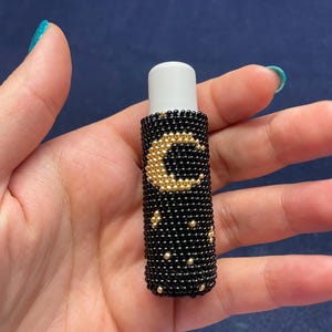 May include: A black beaded lip balm holder with a gold crescent moon and star design. The lip balm tube has a white cap. The holder is cylindrical and appears to be handmade.