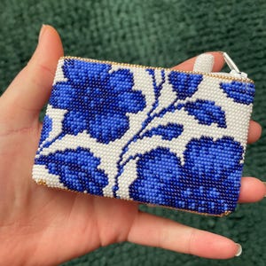 May include: A rectangular beaded coin purse featuring a floral design in blue and white. The purse has a gold-colored zipper and trim. The floral pattern is made of small, tightly packed beads.