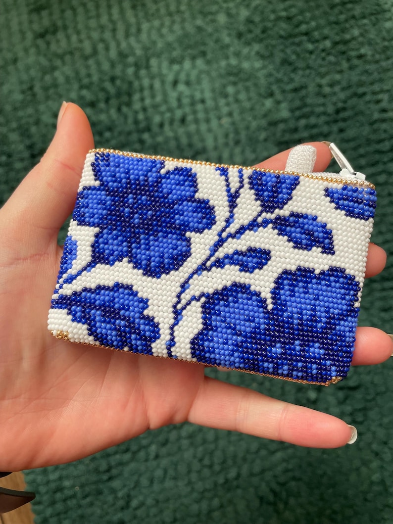 May include: A rectangular beaded coin purse featuring a floral design in blue and white. The purse has a gold-colored zipper and trim. The floral pattern is made of small, tightly packed beads.