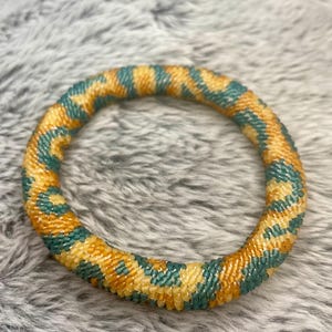 May include: A beaded bracelet with a yellow and teal pattern. The bracelet is made of small, round beads that are woven together in a spiral pattern.