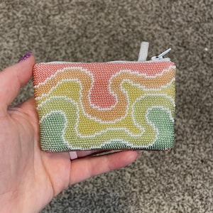 May include: A small, rectangular beaded coin purse with a rainbow color scheme. The purse features a white zipper and a wavy, abstract design in shades of pink, yellow, green, and white. The purse is held in a hand.