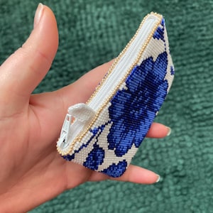 May include: A small, beaded coin purse with a floral design in blue and white. The purse has a white interior and a gold-colored zipper. The edges are trimmed with small gold beads. The purse is held in a hand.