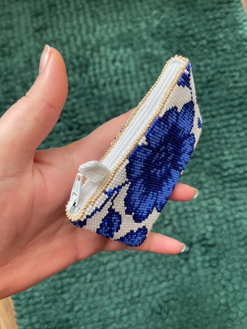 May include: A small, beaded coin purse with a floral design in blue and white. The purse has a white interior and a gold-colored zipper. The edges are trimmed with small gold beads. The purse is held in a hand.