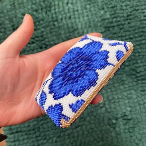 May include: A rectangular beaded clutch featuring a large blue floral design on a white background. The edges are trimmed with gold beads. The clutch is held in a hand.