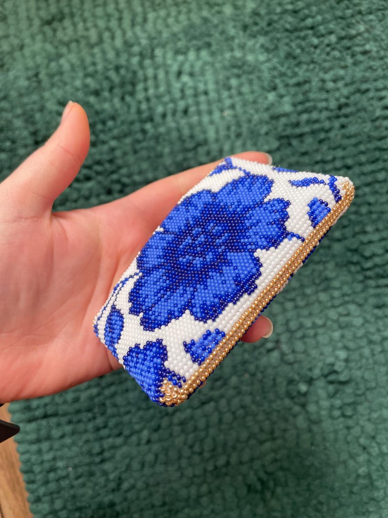 May include: A rectangular beaded clutch featuring a large blue floral design on a white background. The edges are trimmed with gold beads. The clutch is held in a hand.