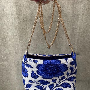 May include: A rectangular handbag with a white background and a blue floral pattern. The bag has a gold chain strap and a black interior. The bag is held up by a hand.