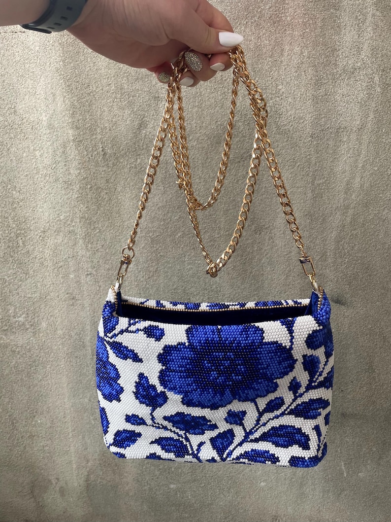 May include: A rectangular handbag with a white background and a blue floral pattern. The bag has a gold chain strap and a black interior. The bag is held up by a hand.