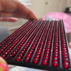 Könnte beinhalten: Nahaufnahme eines roten LED-Displays mit Reihen kleiner, runder, roter Lichter. Das Display ist auf einem schwarzen Rahmen montiert.