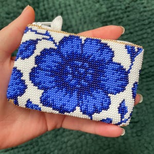 May include: A rectangular beaded coin purse featuring a large blue floral design on a white background. The purse has a gold zipper and trim. The floral pattern is made of small blue beads, creating a textured appearance.