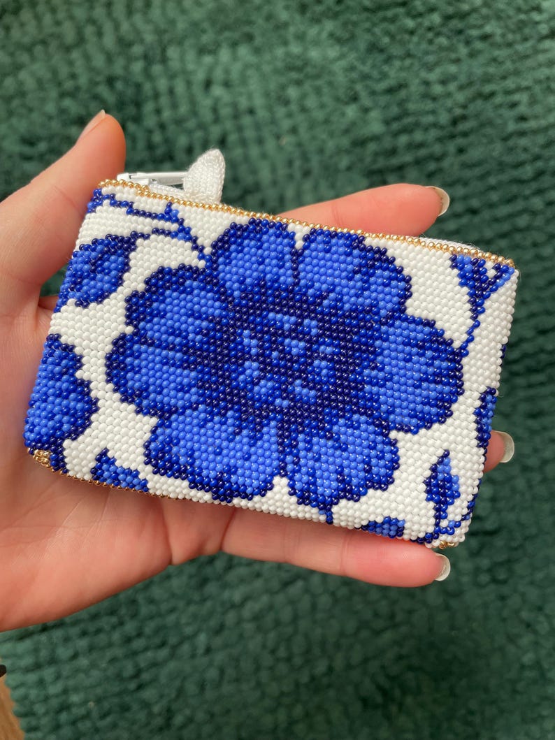 May include: A rectangular beaded coin purse featuring a large blue floral design on a white background. The purse has a gold zipper and trim. The floral pattern is made of small blue beads, creating a textured appearance.