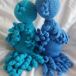 Puede incluir: Dos peluches de pulpo de crochet, uno azul y otro turquesa, con cuerpos y tent&aacute;culos texturizados. Los peluches est&aacute;n sentados sobre una superficie blanca.