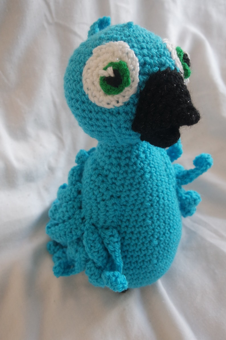 Puede incluir: Un p&aacute;jaro azul de crochet con ojos blancos y verdes y un pico negro. El p&aacute;jaro est&aacute; sentado sobre una superficie blanca.
