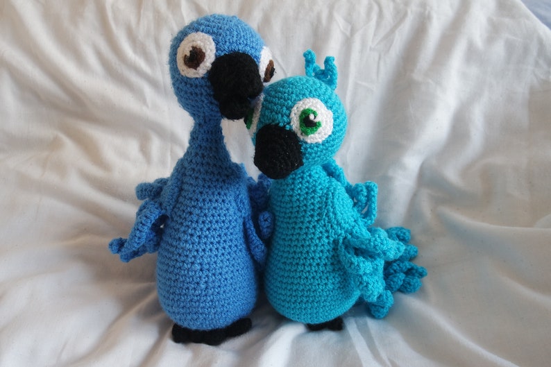Puede incluir: Dos loros de crochet azul y turquesa, uno con ojos marrones y el otro con ojos verdes, de pie uno al lado del otro sobre una superficie blanca.