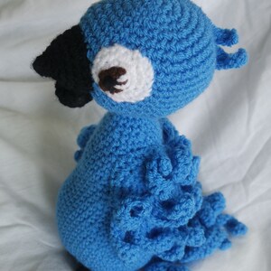 Puede incluir: Un juguete de loro de crochet azul con pico negro y ojo blanco. El loro tiene una cola rizada y est&aacute; sentado sobre una superficie blanca.