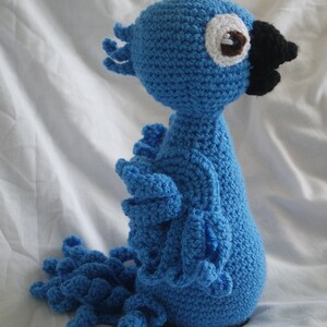 Puede incluir: Juguete de peluche de loro azul tejido a crochet con un ojo blanco y un pico negro. El loro tiene una cola larga con un dise&ntilde;o de volantes.