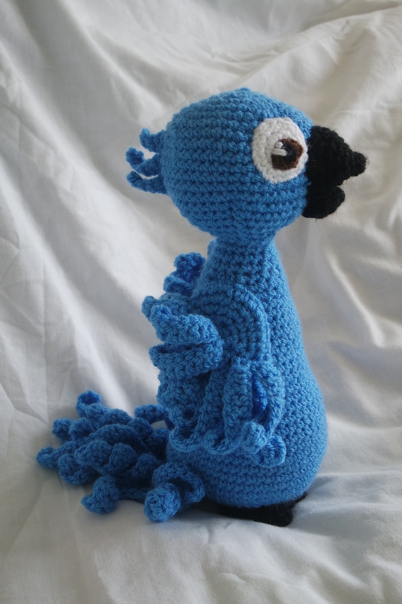 Puede incluir: Juguete de peluche de loro azul tejido a crochet con un ojo blanco y un pico negro. El loro tiene una cola larga con un dise&ntilde;o de volantes.