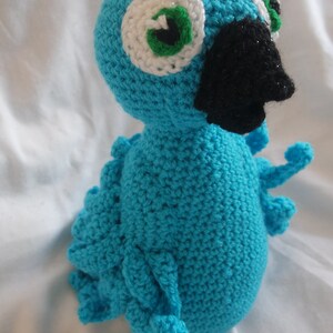 Puede incluir: Un p&aacute;jaro azul de crochet con ojos blancos y verdes y un pico negro. El p&aacute;jaro est&aacute; sentado sobre una superficie blanca.