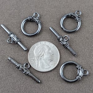 May include: Three dark metallic toggle clasps, each consisting of a circular ring and a bar component with decorative textured detailing. A United States quarter coin is placed in the center for scale, measuring approximately 0.95 inches in diameter.