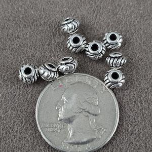 May include: Close-up of silver-colored beads with a detailed, textured design. The beads are scattered on a gray surface next to a U.S. quarter coin. The beads appear to be approximately 0.25 inches in diameter.