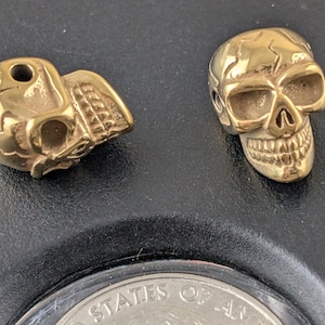 May include: Two small, gold-colored skull beads. One skull is shown in profile, and the other is facing forward. The skulls have detailed features, including eye sockets, a nose cavity, and teeth. The beads are likely for jewelry making.