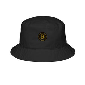 May include: A solid black bucket hat featuring a circular patch centred on the front. The patch displays a gold-coloured Bitcoin symbol set against a dark background, with a textured, distressed appearance within the circular border.