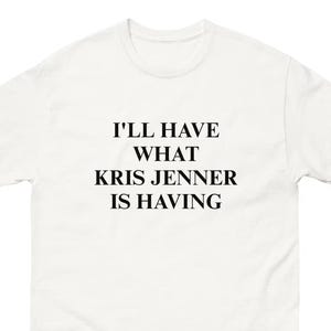 Può includere: T-shirt bianco con la scritta nera "I'LL HAVE WHAT KRIS JENNER IS HAVING" centrata sul davanti. La maglietta ha un girocollo e maniche corte, adatta per un abbigliamento casual.