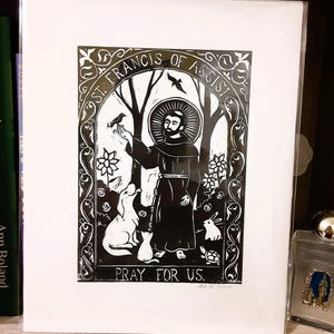 May include: Black and white woodcut print of Saint Francis of Assisi. The print depicts Saint Francis surrounded by animals, including a bird, a dog, a cat, and a rabbit. The text "Pray for Us" is at the bottom of the print.