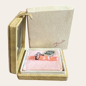 May include: A square, cream-colored textured jewelry box with a hinged lid featuring the brand name Dorette in red script. The interior is lined with a soft, pale pink fabric, a matching pink ribbon, and a small silver-toned metal tag.