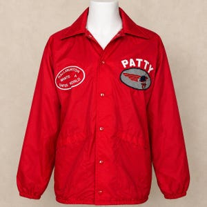 Può includere: Una giacca coach rosso brillante a maniche lunghe con colletto a punta e chiusura frontale con bottoni a pressione. Il petto presenta due toppe: una ovale con la scritta Halliburton Wants A Safer World e l'altra con il nome Patty sopra un logo alato.