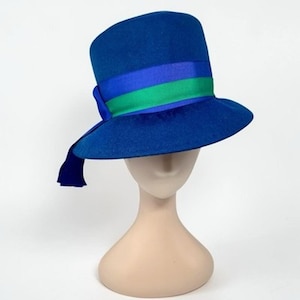 May include: A royal blue felt hat with a flat top and a wide brim. The hat features a decorative band around the crown consisting of a bright blue ribbon layered with a vibrant green ribbon, ending in a short, dangling blue ribbon tail on the side.