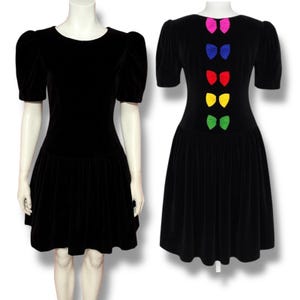 May include: A black velvet dress with short, puffed sleeves and a flared skirt. The back of the dress features a series of colorful bows in pink, blue, red, yellow, and green. The dress is displayed on a mannequin.
