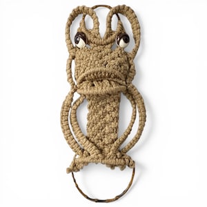May include: A tan macrame wall hanging featuring a stylized frog-like figure with two large, round, dark brown and white eyes. The piece is crafted from knotted cord and includes a circular wooden ring at the bottom for hanging or display purposes.