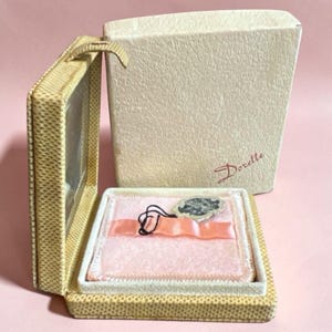 May include: A beige textured jewelry box with a hinged lid and a cream-colored outer box featuring the word Dorette in red script. The interior is lined with light pink fabric, a peach-colored ribbon, and includes a small decorative tag.