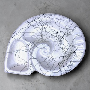 May include: White, shell-shaped serving dish with a black, abstract, vein-like pattern. The dish has three compartments and a spiral design in the center. The dish is on a gray surface.