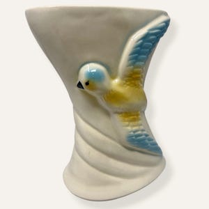 May include: A white ceramic vase featuring a blue and yellow bird motif. The bird is depicted mid-flight, with its wings spread. The vase has a curved form and a textured, swirling design.