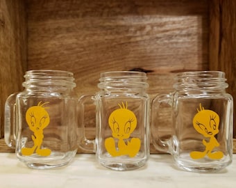 May include: Three clear glass mason jars with yellow handles. Each jar has a yellow cartoon image of Tweety Bird on the front.