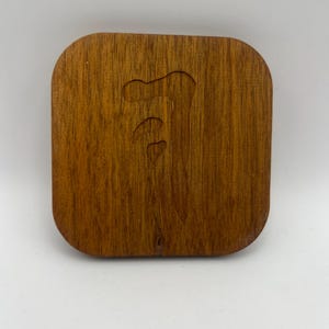 May include: A square wooden lid with rounded corners. The lid is a warm brown color with a visible wood grain. A carved design is visible on the surface, featuring a series of curved shapes. The lid appears to be for a small box or container.