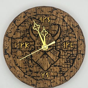 May include: Circular wooden clock with a carved, textured surface. The clock face features gold-coloured hands and Roman numerals. A bull's head is carved in the centre. The clock has a rustic, antique aesthetic.