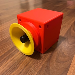 May include: A red cube with a yellow and black speaker attached to one side. The speaker has a small black knob in the center.