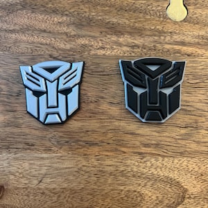 May include: Two 3D printed Transformers logos, one in white and one in black, on a wooden surface.