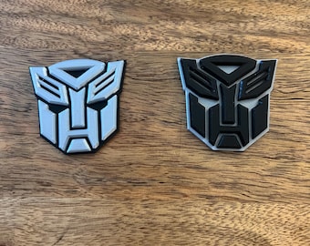 May include: Two 3D printed Transformers logos, one in white and one in black, on a wooden surface.