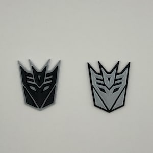Op de afbeelding: Twee 3D-geprinte versies van het Decepticons-logo. Een is zwart en de andere is zilver.
