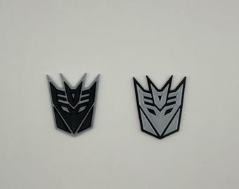 May include: Two 3D printed versions of the Decepticons logo. One is black and the other is silver.