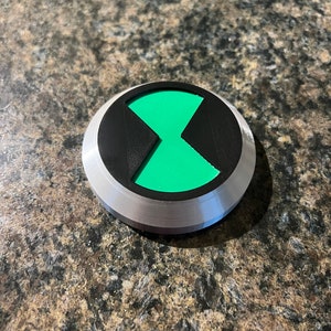 May include: A silver and black circular button with a green hourglass symbol in the center.